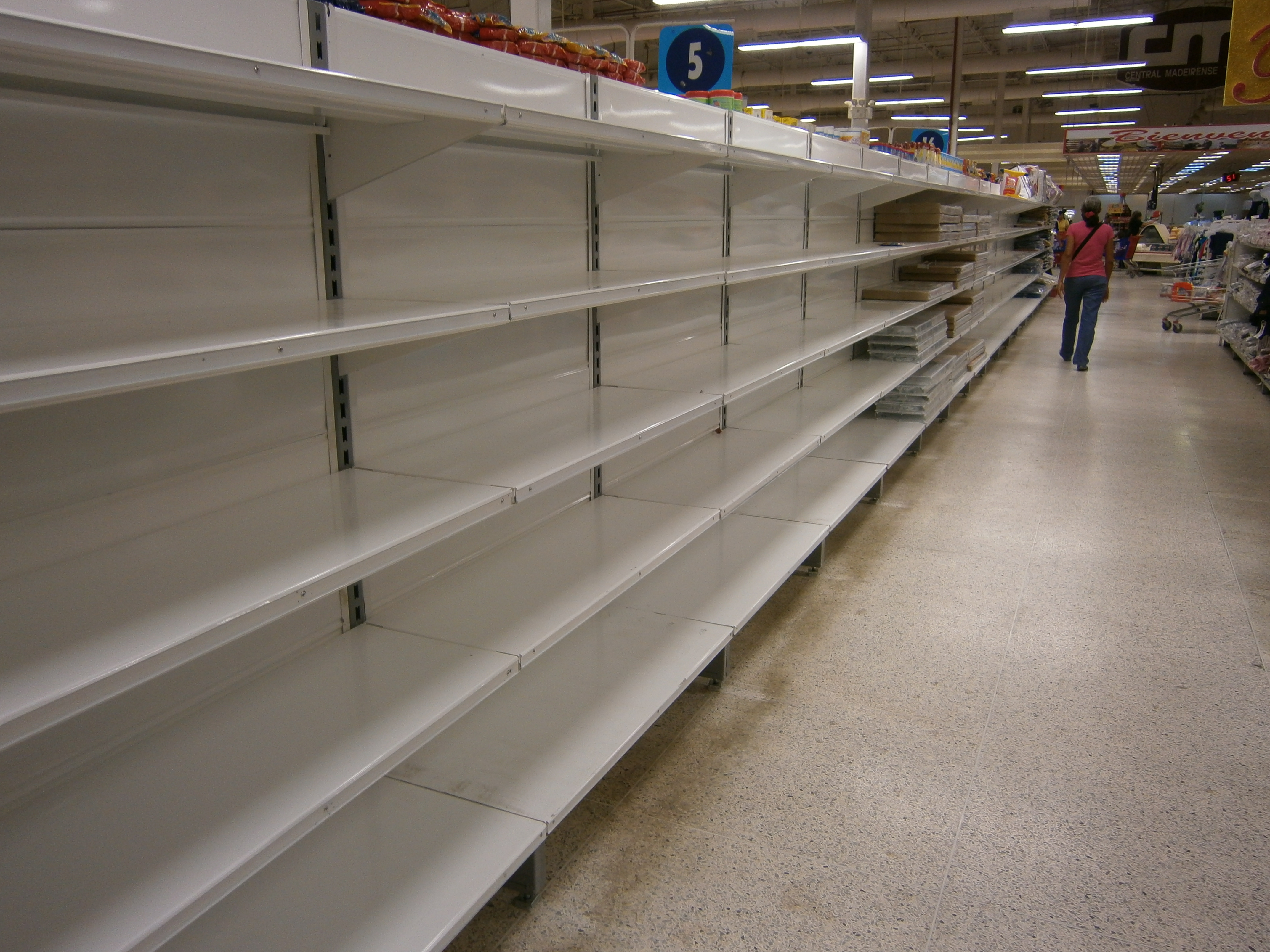 Mostly empty supermarket shelves in Venezuela with a few shoppers visible in the distance.
